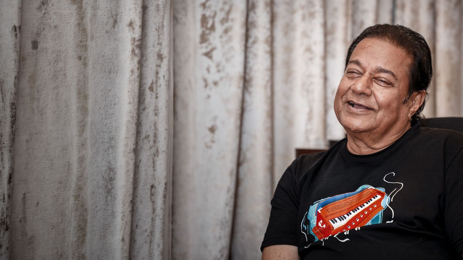 Smiling middle-aged man in a black graphic t-shirt sitting relaxed against a curtained background.
