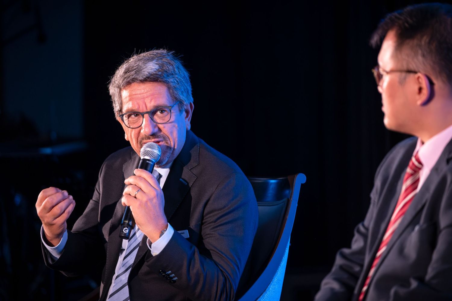 A man in a dark blazer speaks into a microphone during a panel discussion at a professional conference.