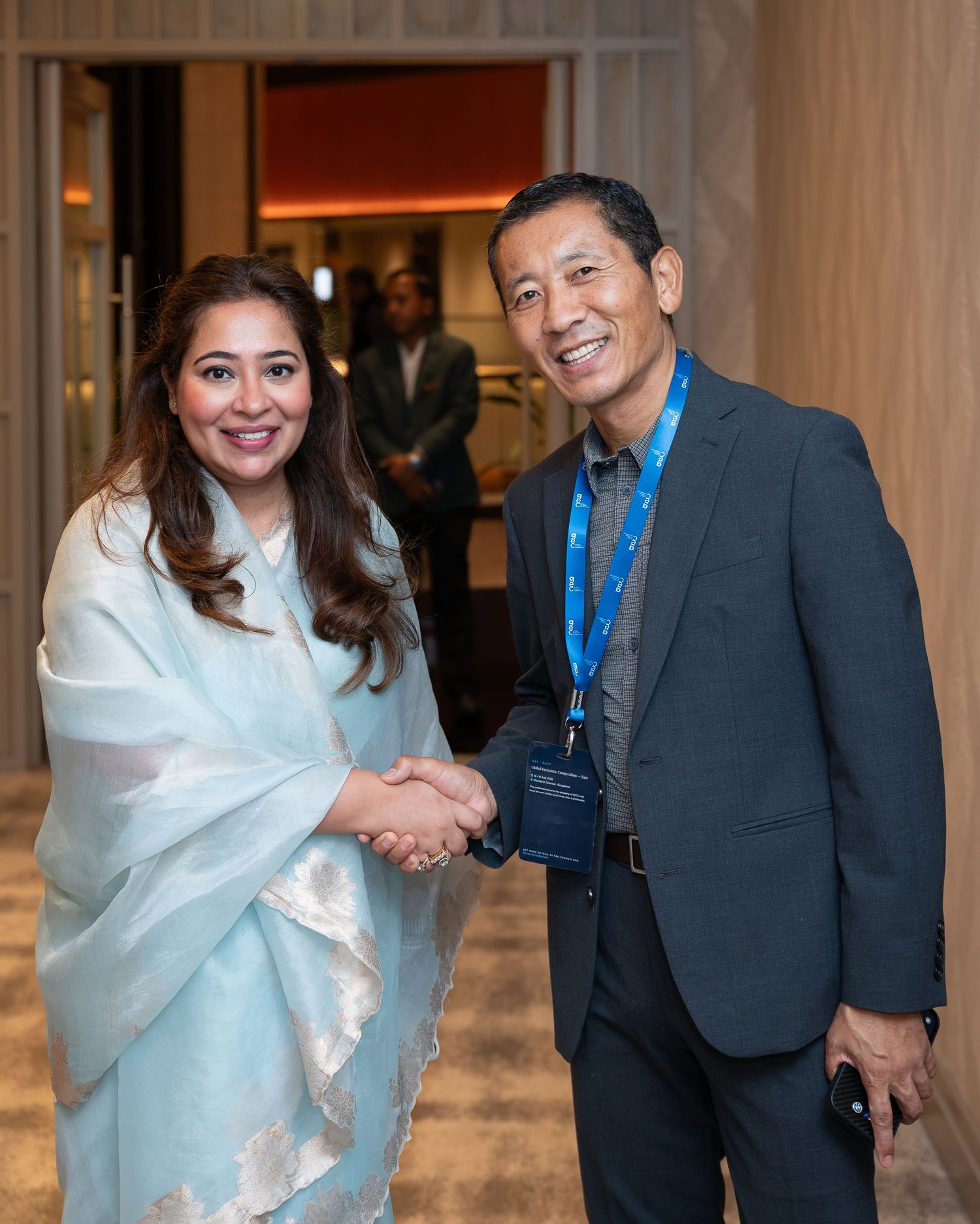 Two professionals shaking hands and smiling at a formal networking event, woman in light blue shawl and man in dark suit.