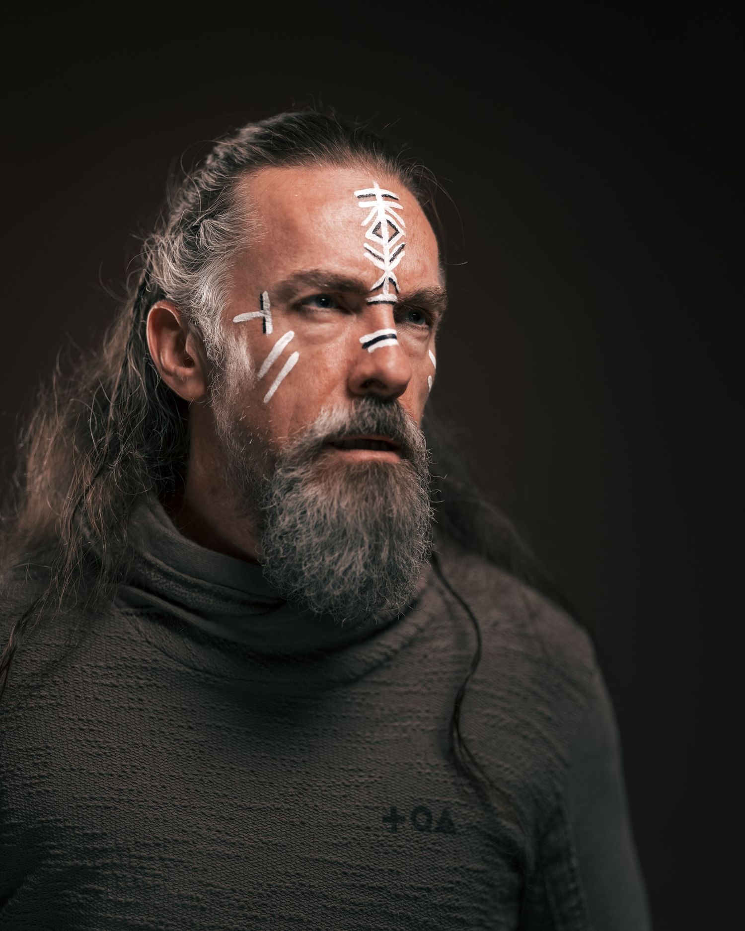 Bearded man with long hair and white tribal face paint wearing a dark turtleneck, dramatic studio portrait.