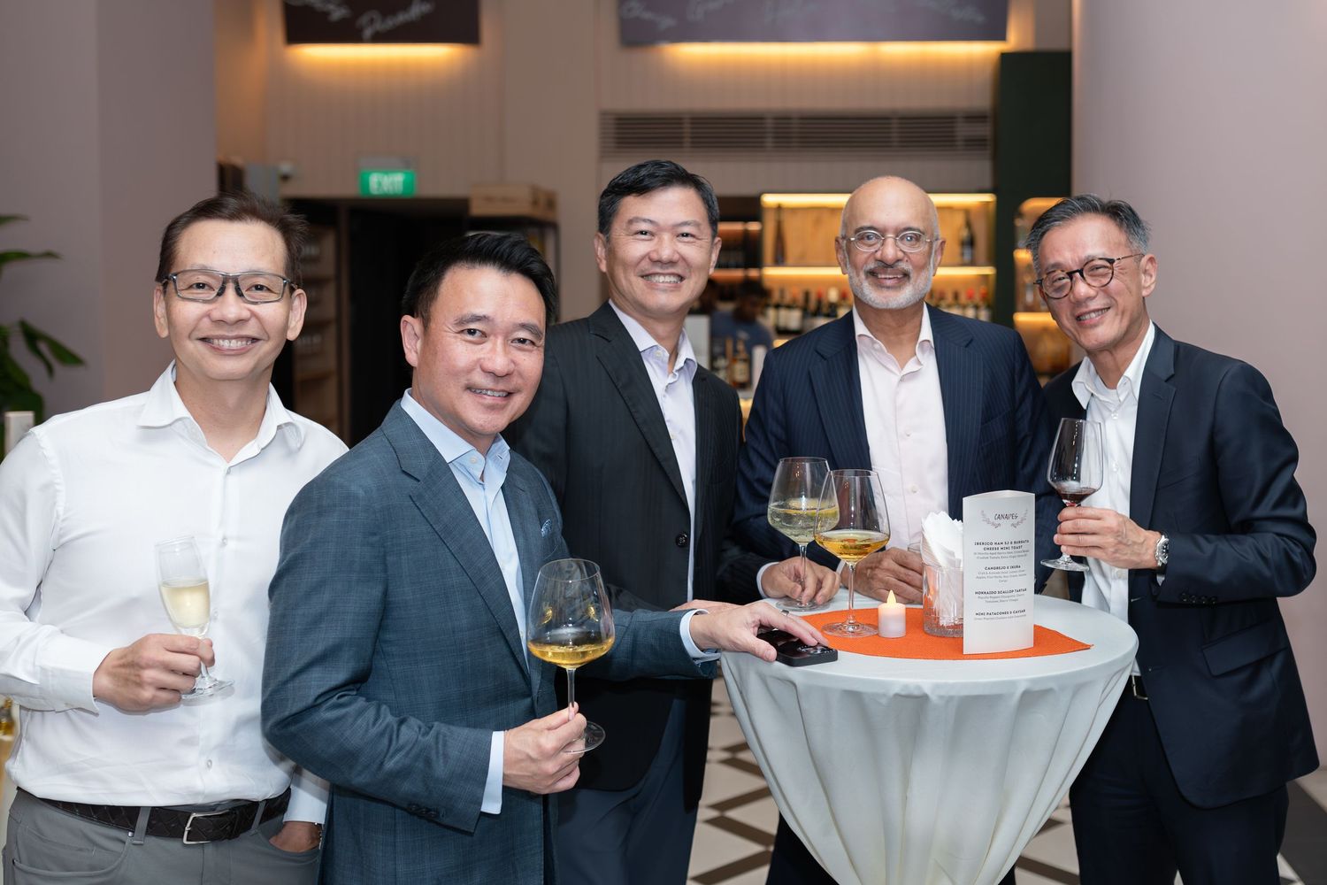 Five men socializing at an elegant event, holding wine glasses around a high-top table.