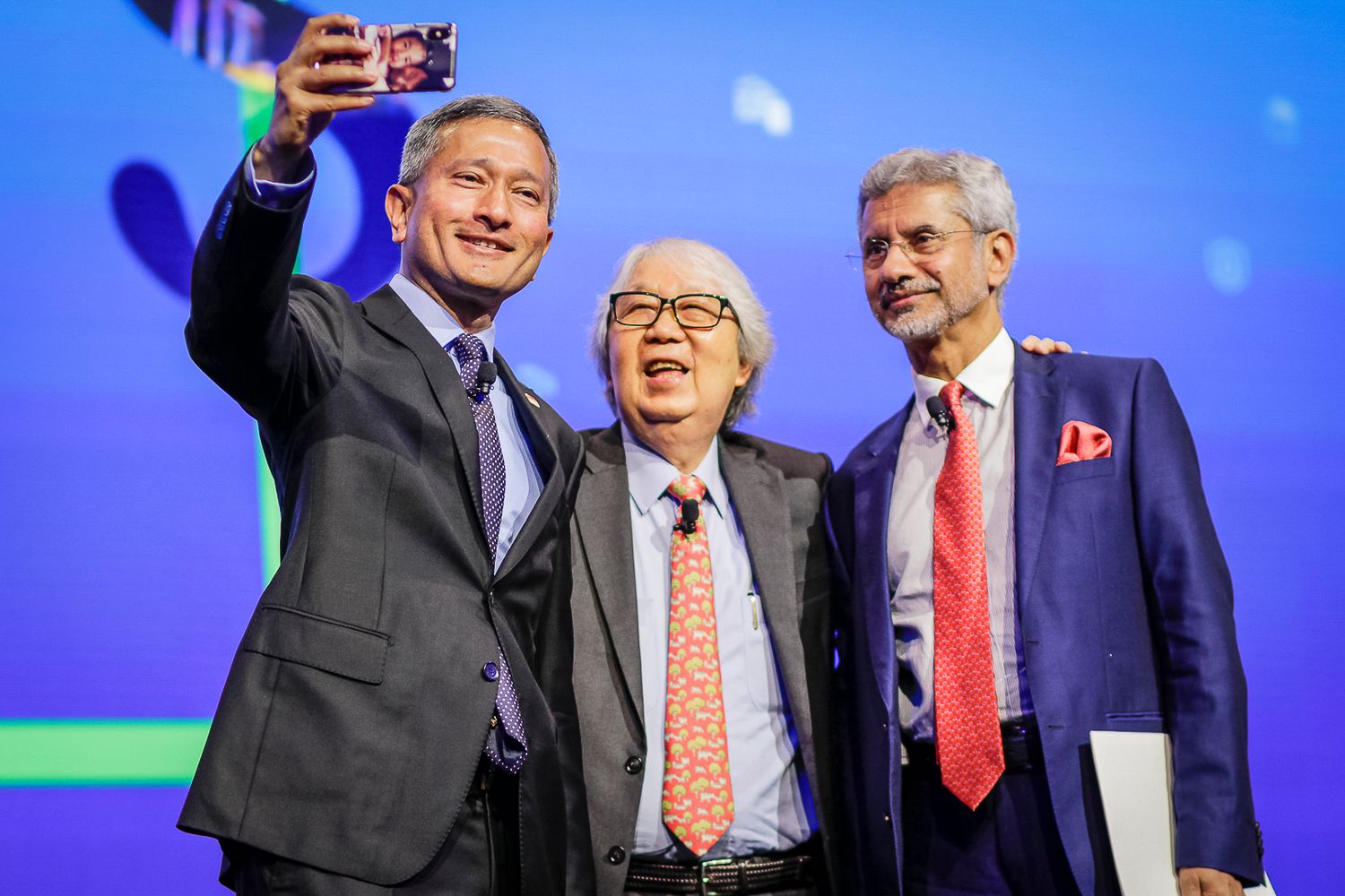 Three men take a selfie together at a formal conference or diplomatic event on stage.