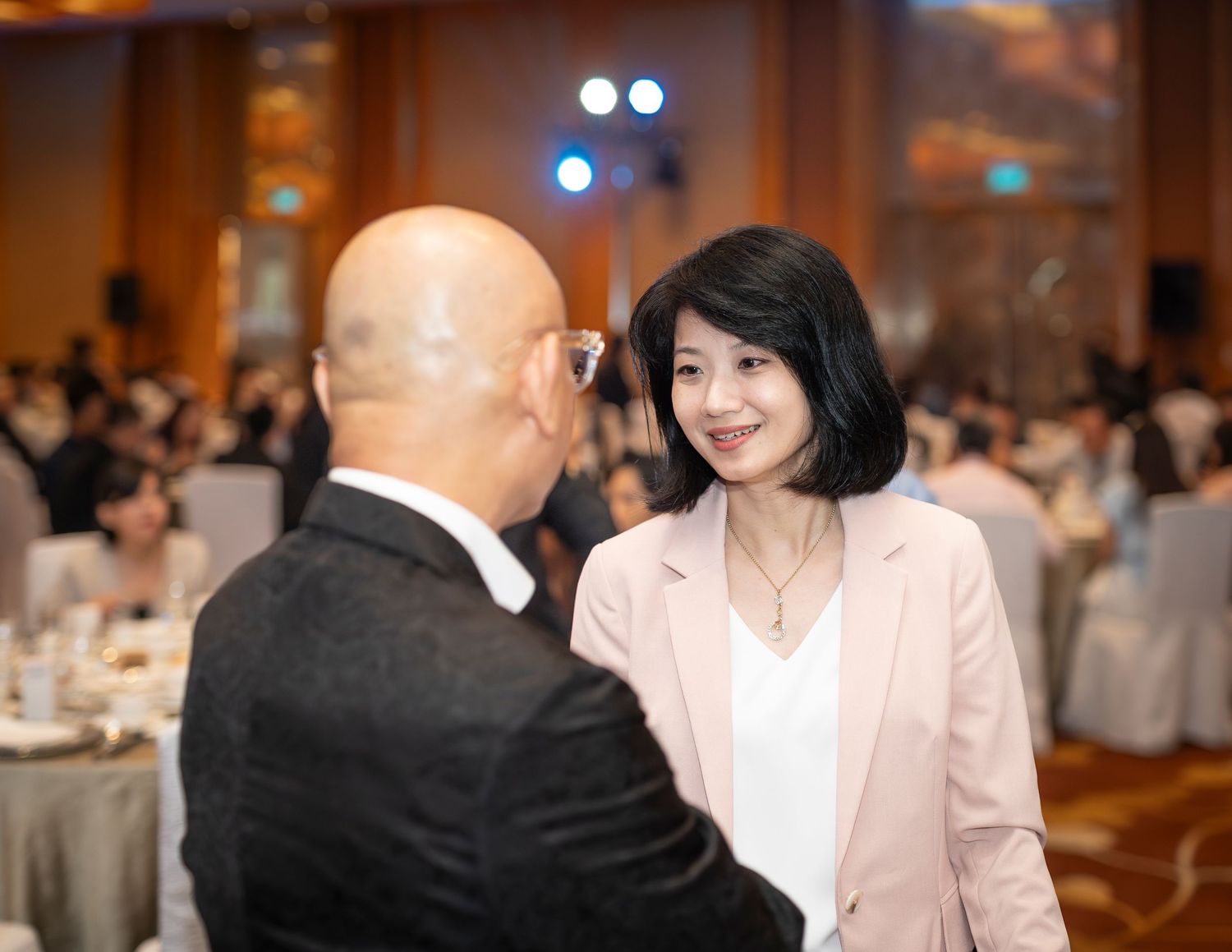 Woman in pink blazer converses with bald man at formal gala dinner event.