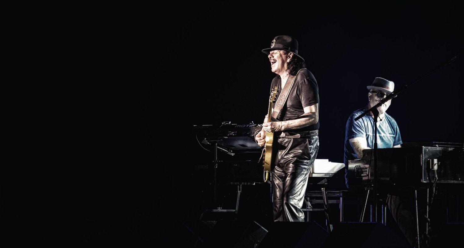 Two musicians performing on a darkened stage, one playing electric guitar and another at a keyboard.