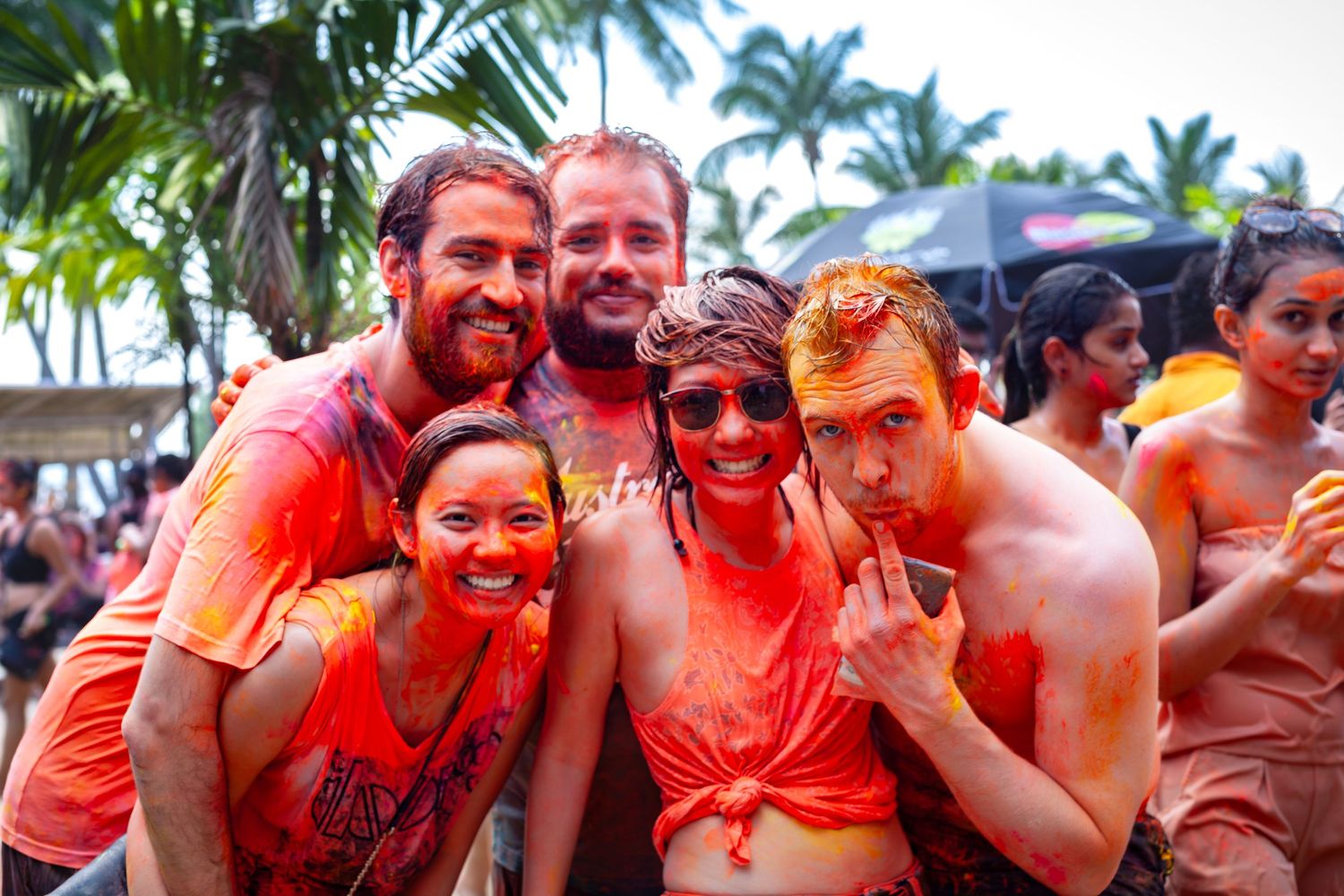 Group of smiling friends covered in colorful powder paint at an outdoor Holi festival event.