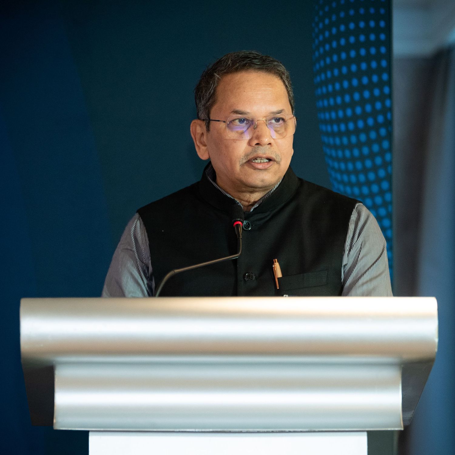 Man in black vest speaking at a silver podium during a formal conference event.