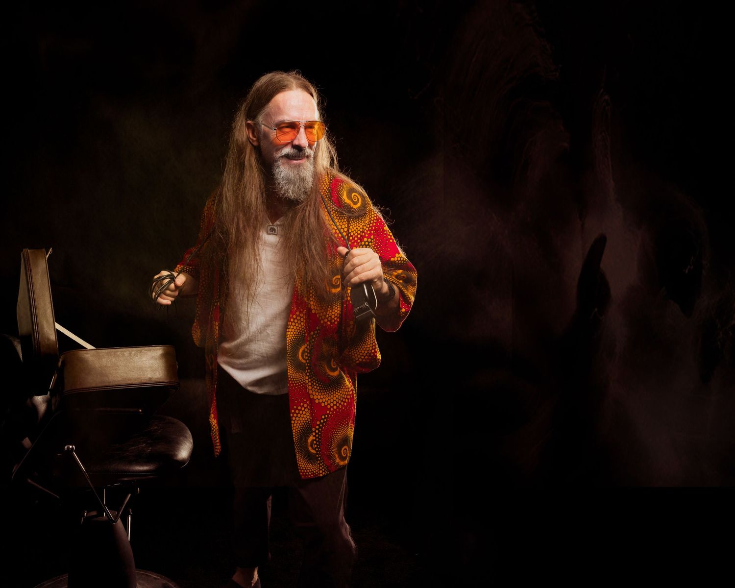 Elderly man with long gray hair and beard wearing colorful psychedelic jacket and orange sunglasses on dark stage.