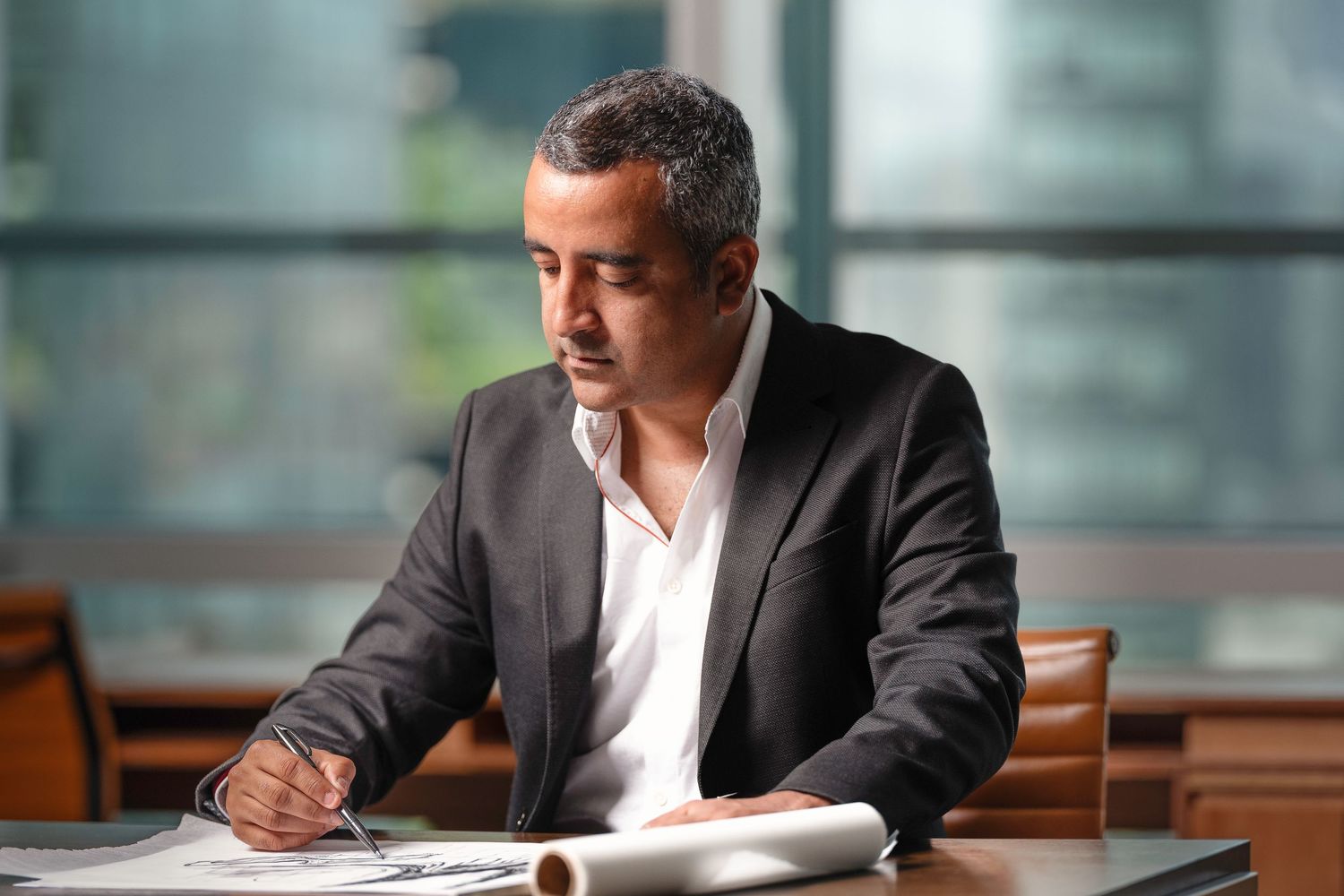 Professional businessman in black blazer writing at desk in modern office with city view.