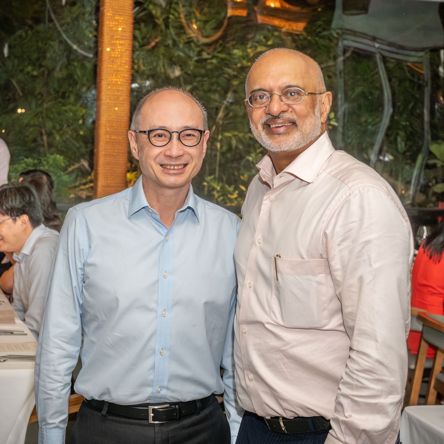 Two men smiling and posing together at an outdoor dining event surrounded by lush greenery.