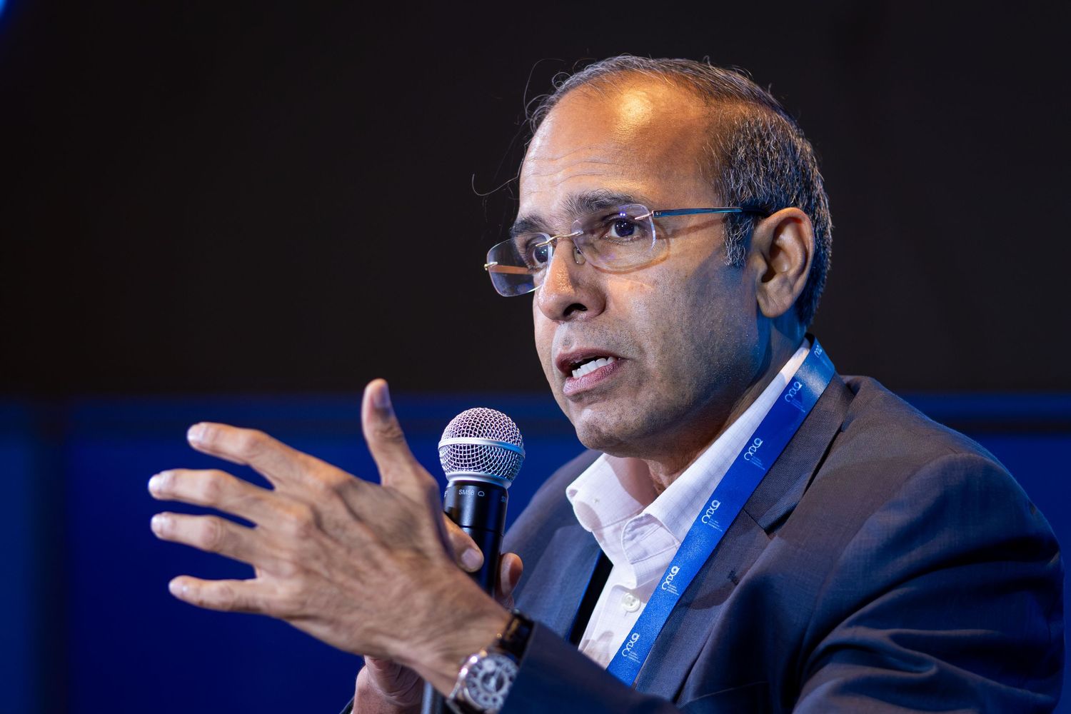 Man in suit speaking at conference panel gesturing with hands holding microphone.