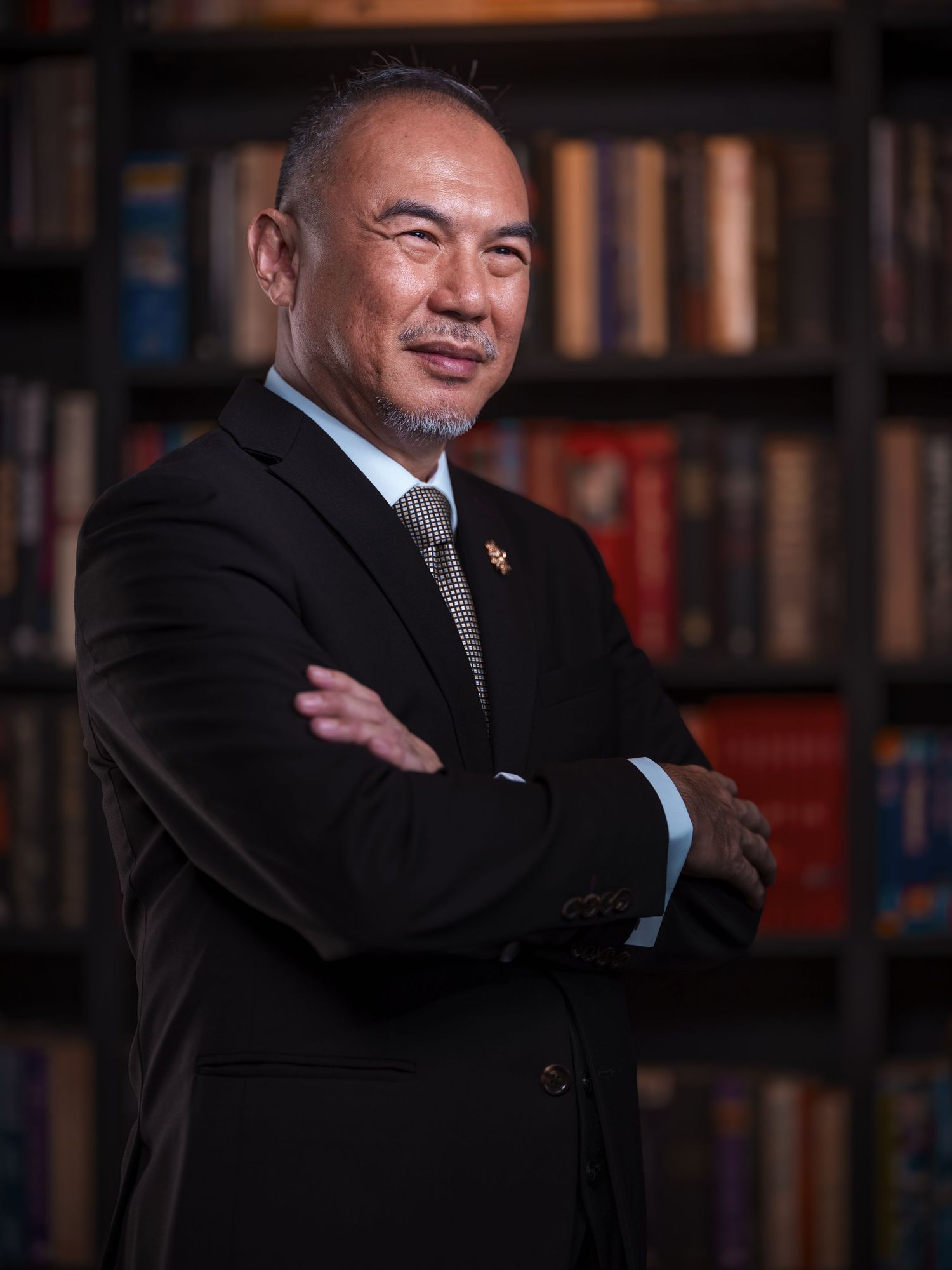 Professional man in black suit with crossed arms standing before a bookshelf library background.