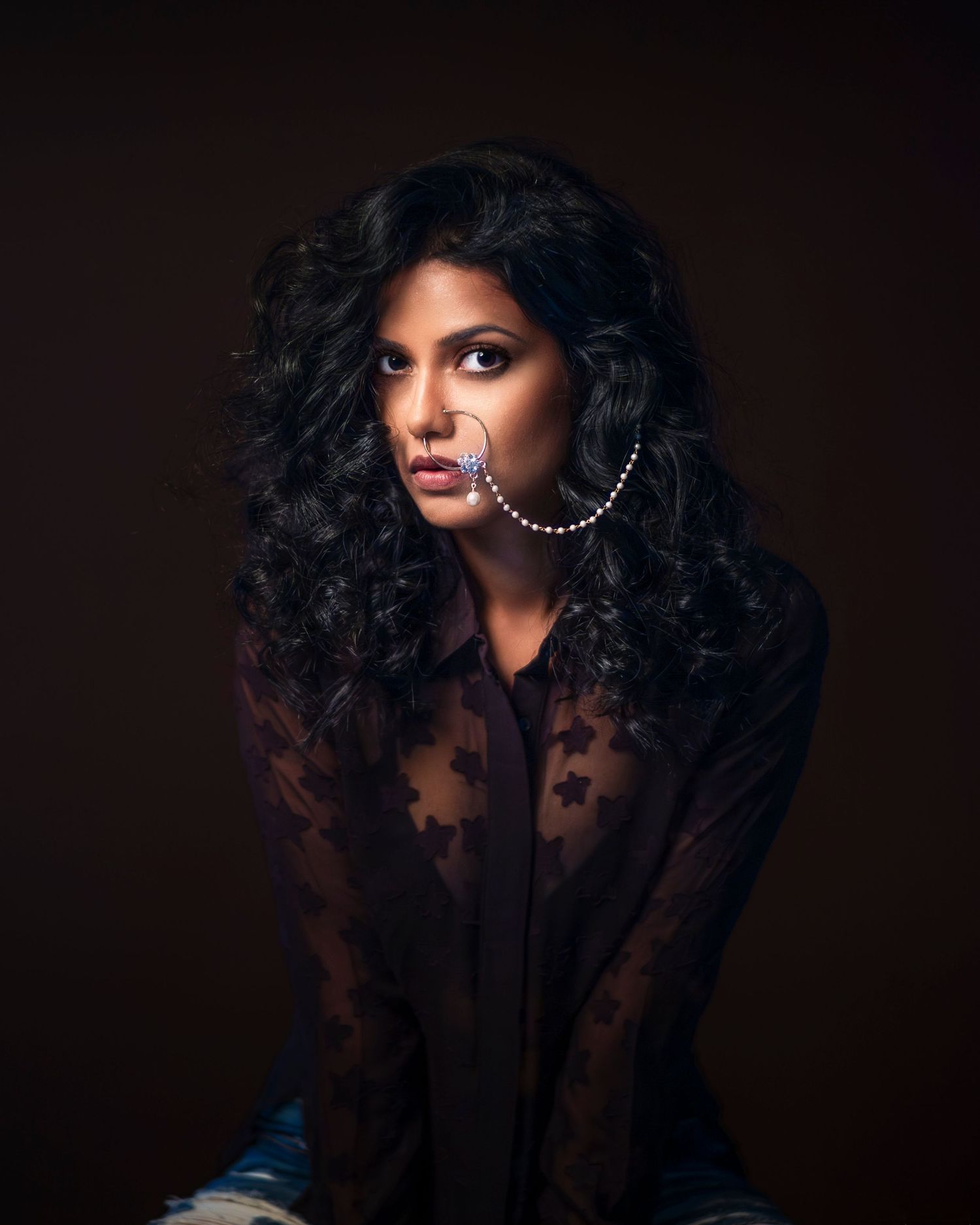Woman with curly black hair wearing sheer lace blouse and hoop earrings posing against dark background.
