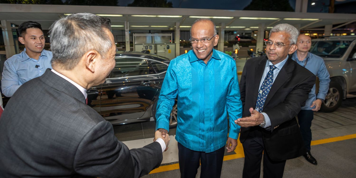 Three men in formal attire shaking hands at a car dealership showroom during an official visit.