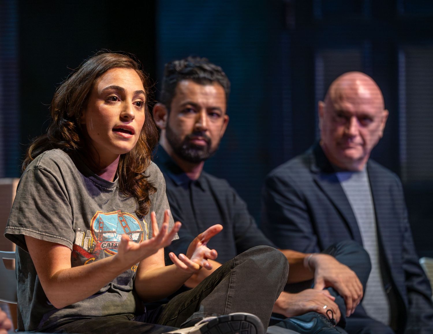 Young woman speaks passionately during a panel discussion with two men seated behind her on stage.