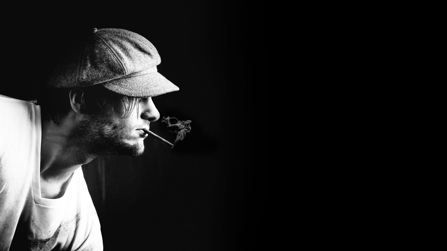 Black and white portrait of man wearing cap smoking pipe against dark background.