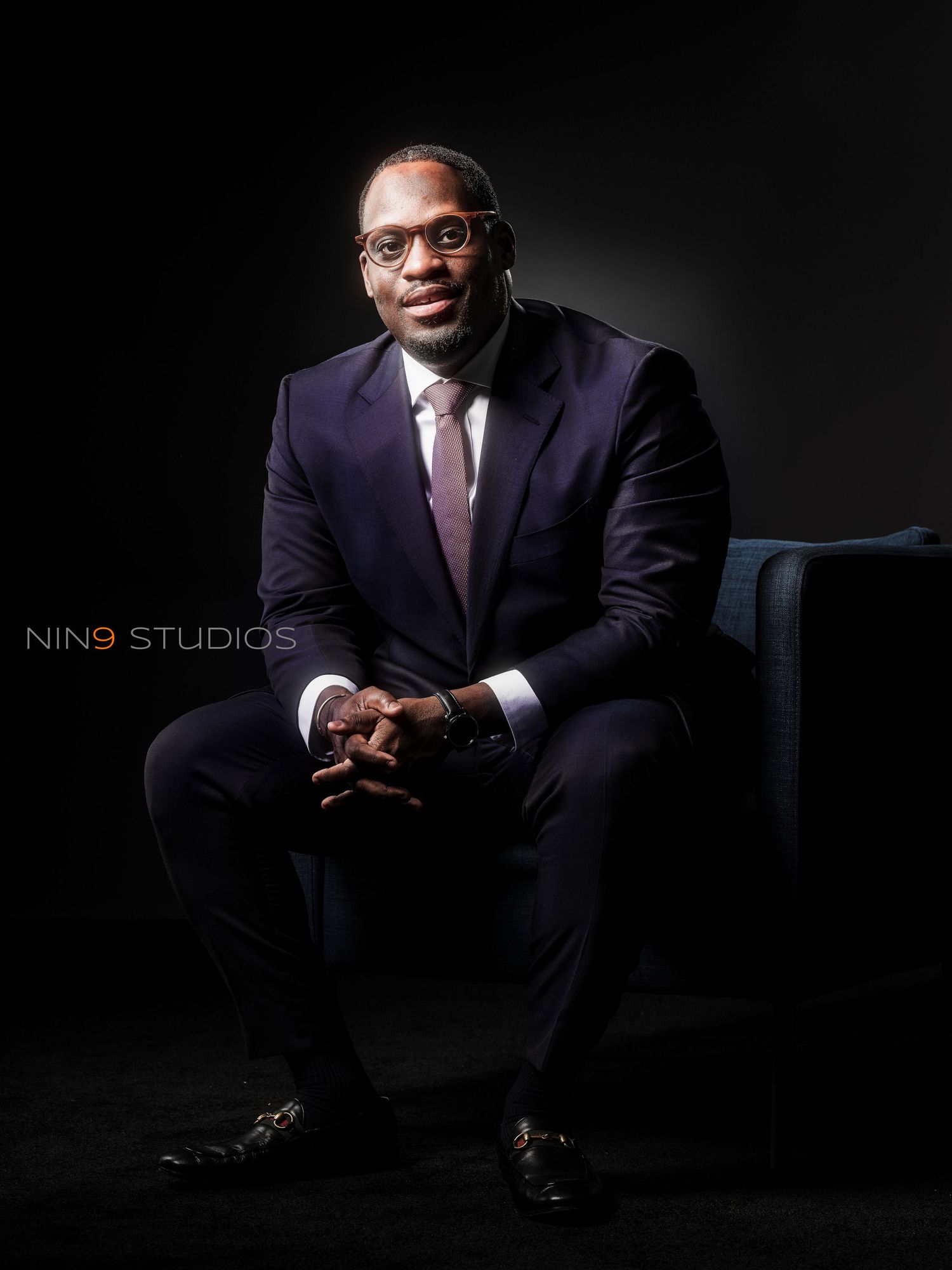 Professional man in navy suit and pink tie seated confidently in studio portrait by Nin9 Studios.