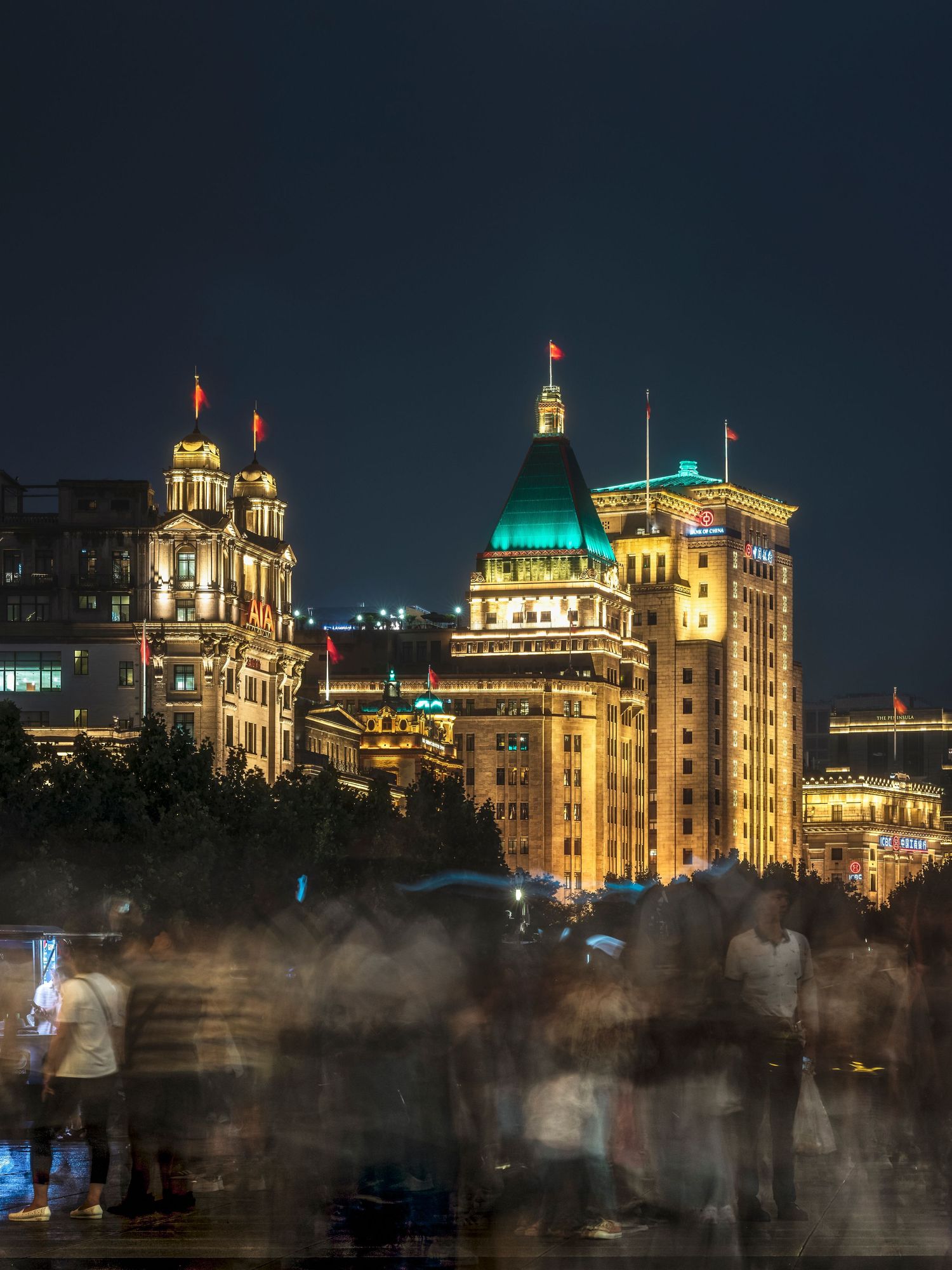 Illuminated historic buildings of the Bund in Shanghai at night with blurred crowds in the foreground.