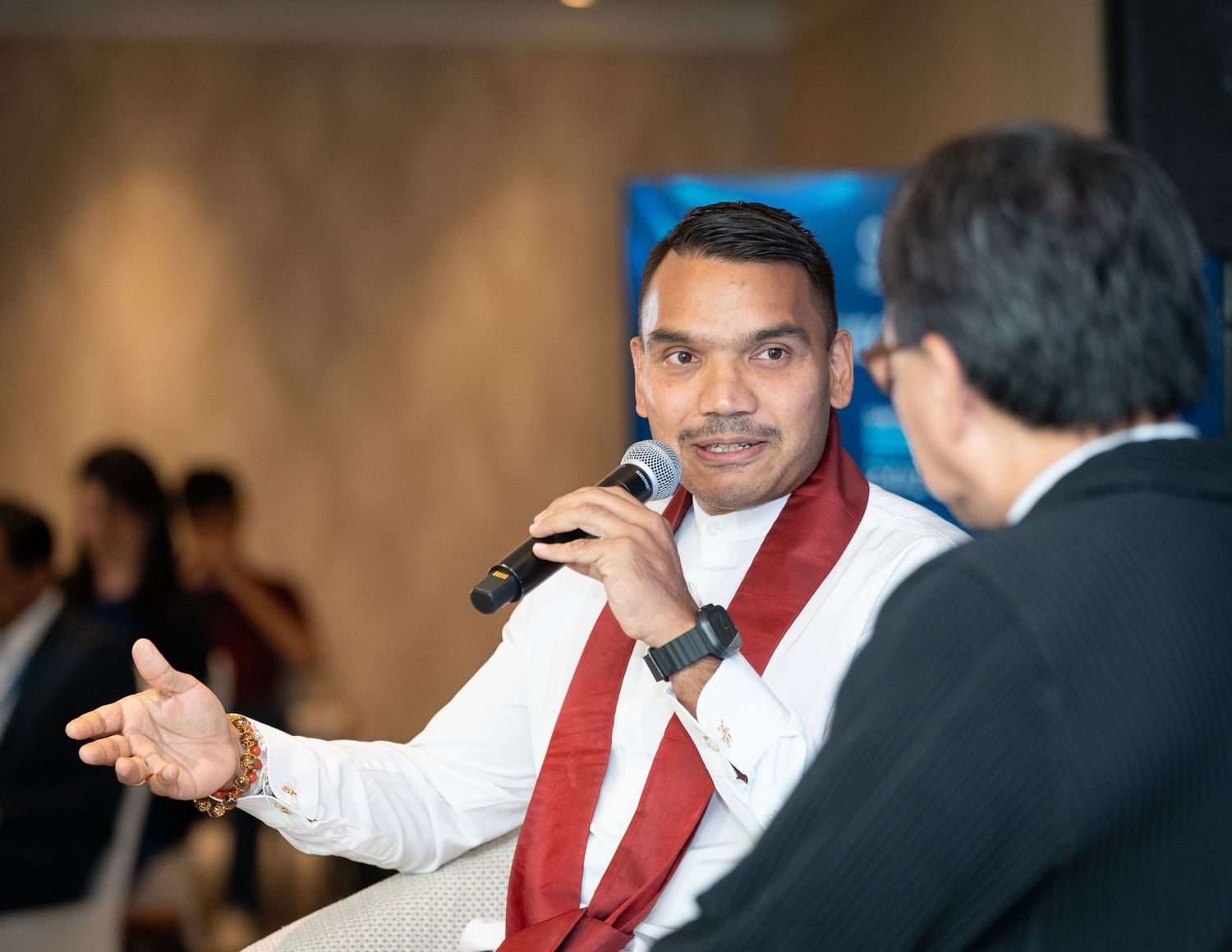 Man in traditional Sri Lankan attire speaks into microphone at conference panel event.