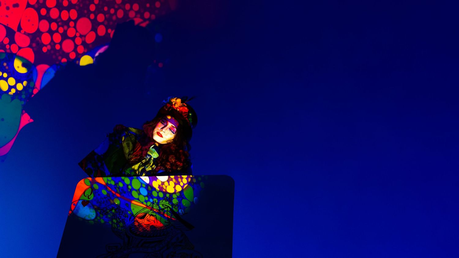 A performer in colorful costume glows under neon UV blacklight with vibrant paint splashes in a dark blue setting.