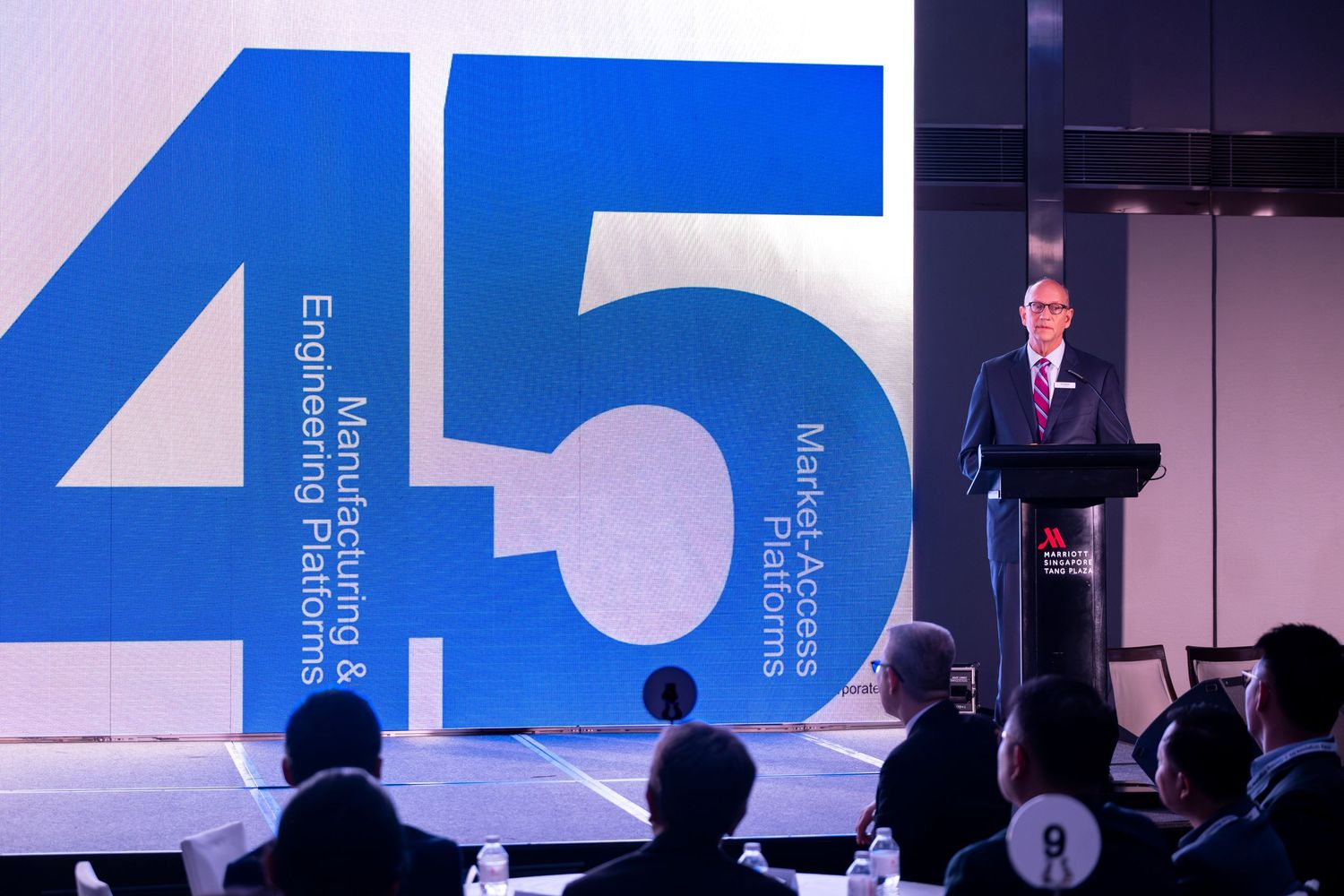 Business executive speaking at a podium in front of a large blue sign displaying the number 45 at a corporate event.