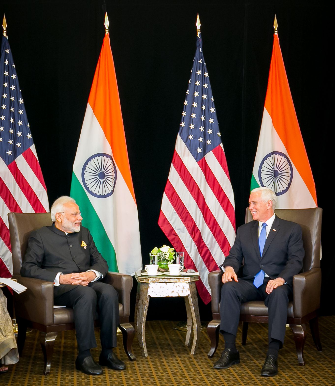 Two leaders sit in chairs flanked by Indian and American flags during a formal bilateral diplomatic meeting.