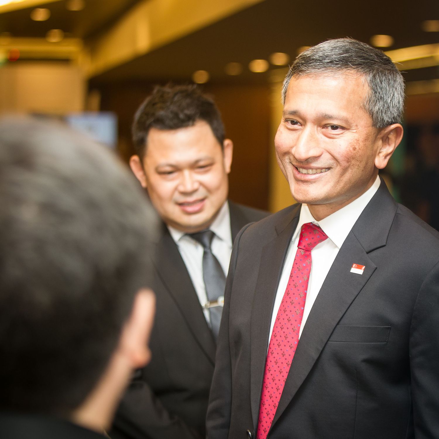 A smiling diplomat in a dark suit with Singapore flag pin engages in conversation at a formal reception.