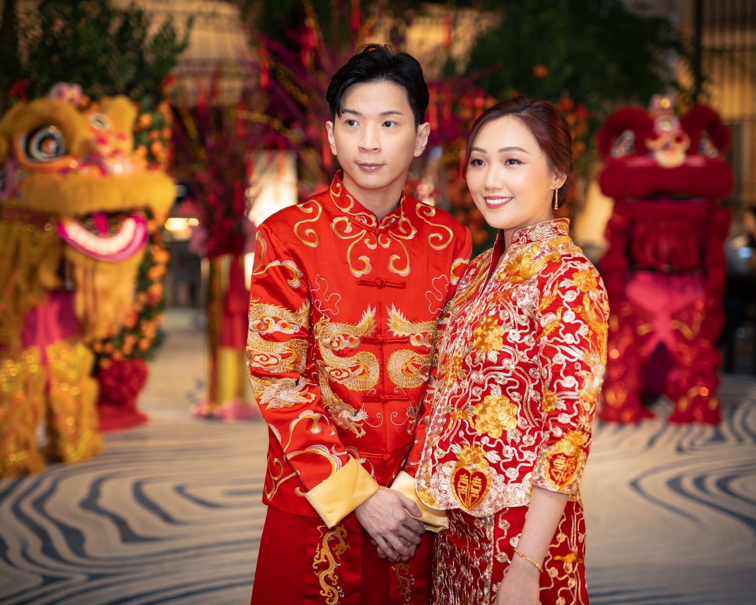 Couple in traditional red Chinese wedding attire with dragon embroidery, lion dance backdrop.