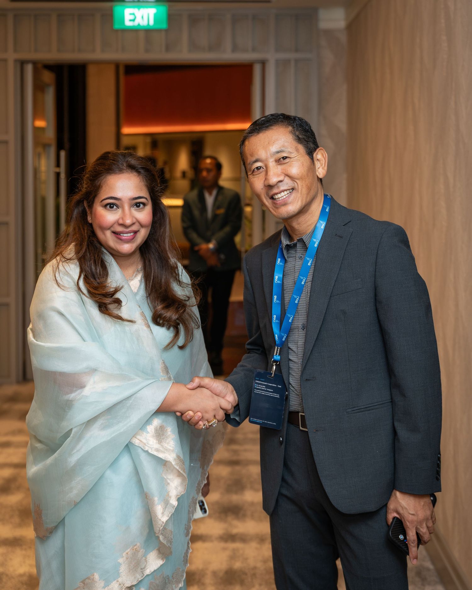 A woman in a light blue saree shakes hands with a man in a dark suit wearing a blue lanyard at a formal event.