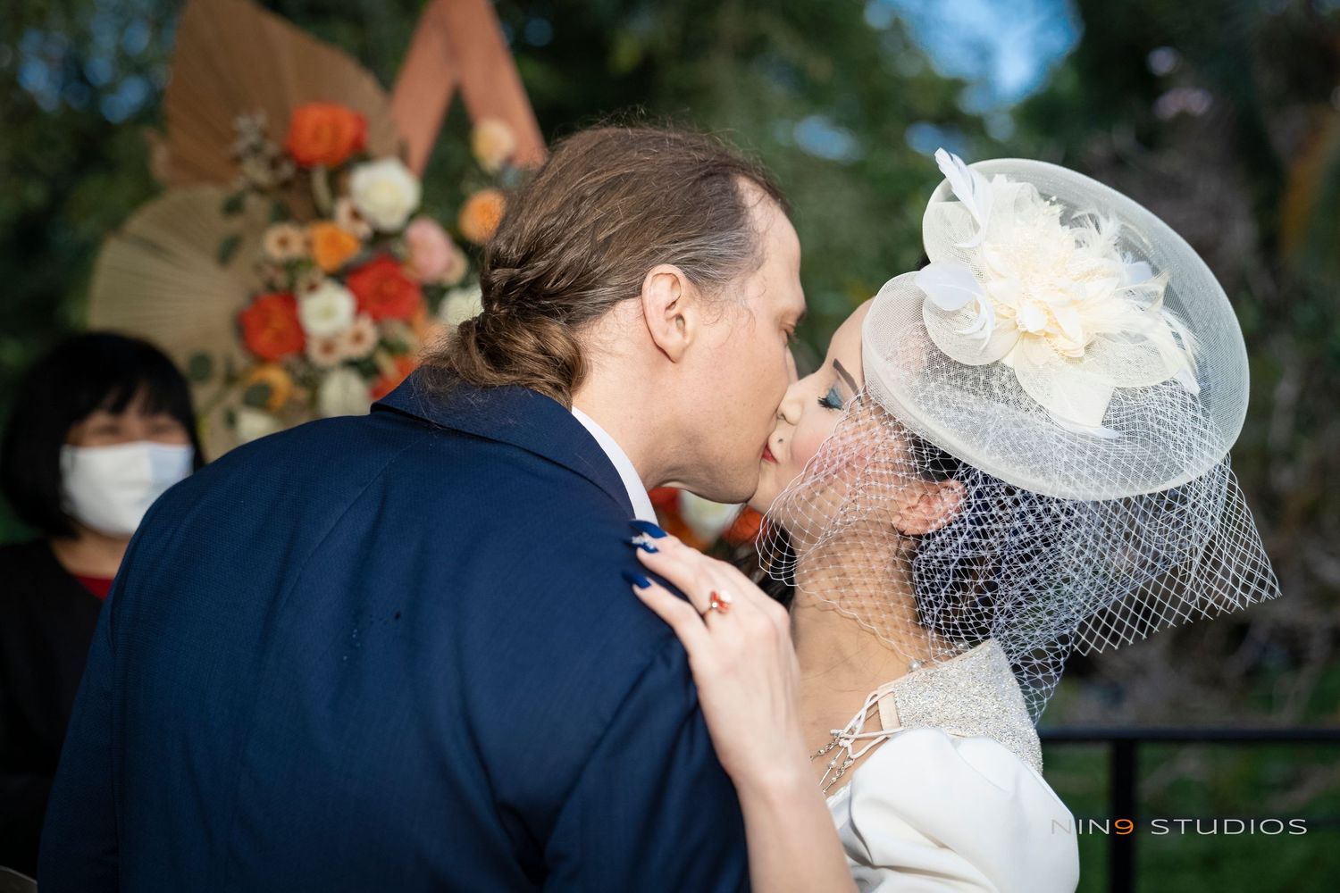 A bride in a white fascinator hat and groom in a navy suit share a kiss at an outdoor wedding ceremony.