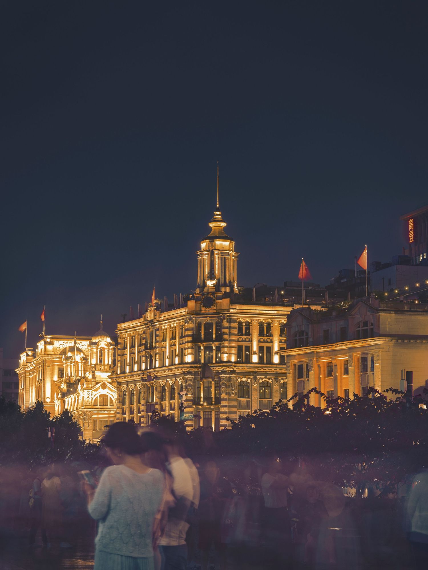 Illuminated historic buildings along the Bund in Shanghai at night with crowds of tourists in the foreground.