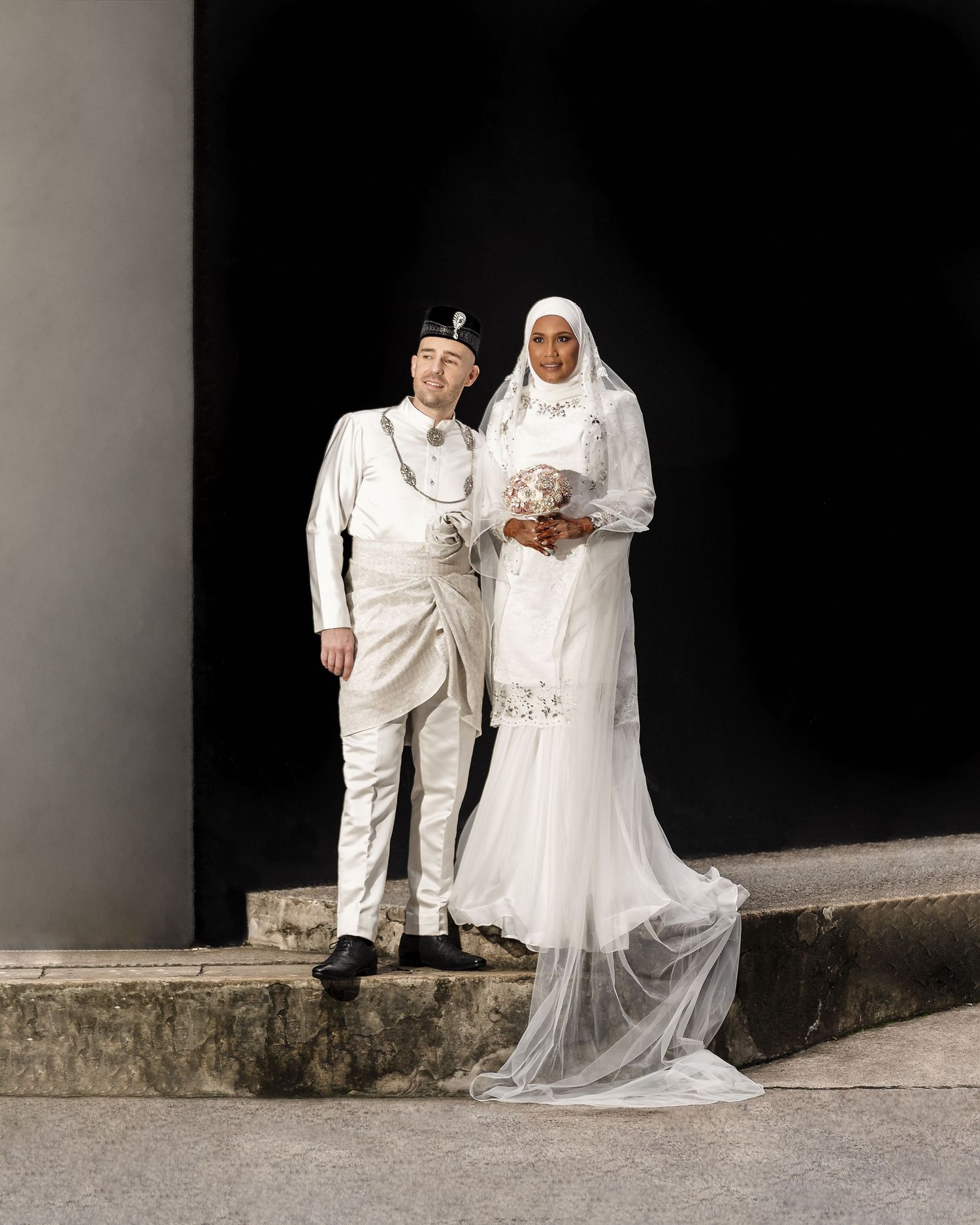 A couple in traditional Malay wedding attire, groom in white baju melayu and bride in white hijab gown, posing outdoors.