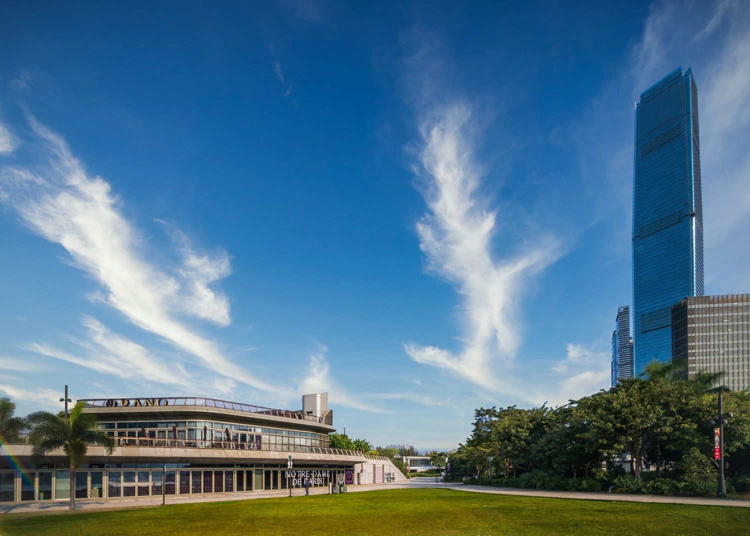Modern waterfront building with lush greenery and a tall glass skyscraper under a vibrant blue sky with wispy clouds.