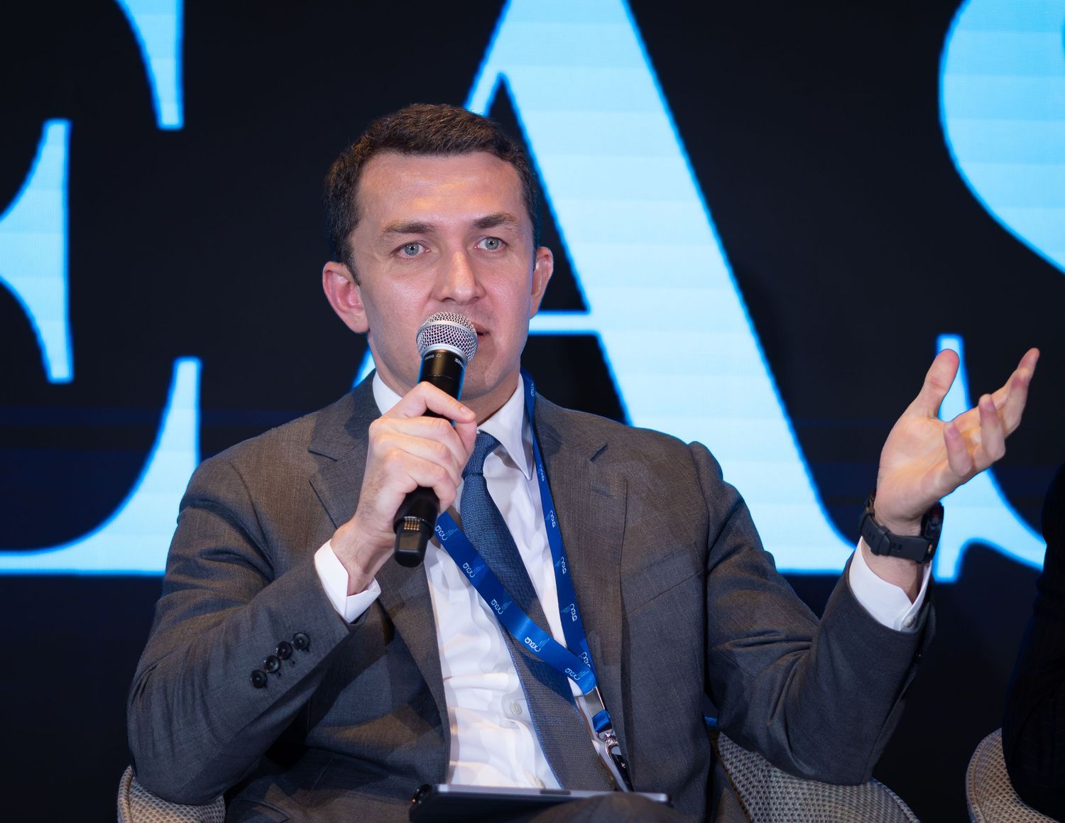 Man in gray suit speaking at a conference panel, holding microphone with a blue lanyard badge.