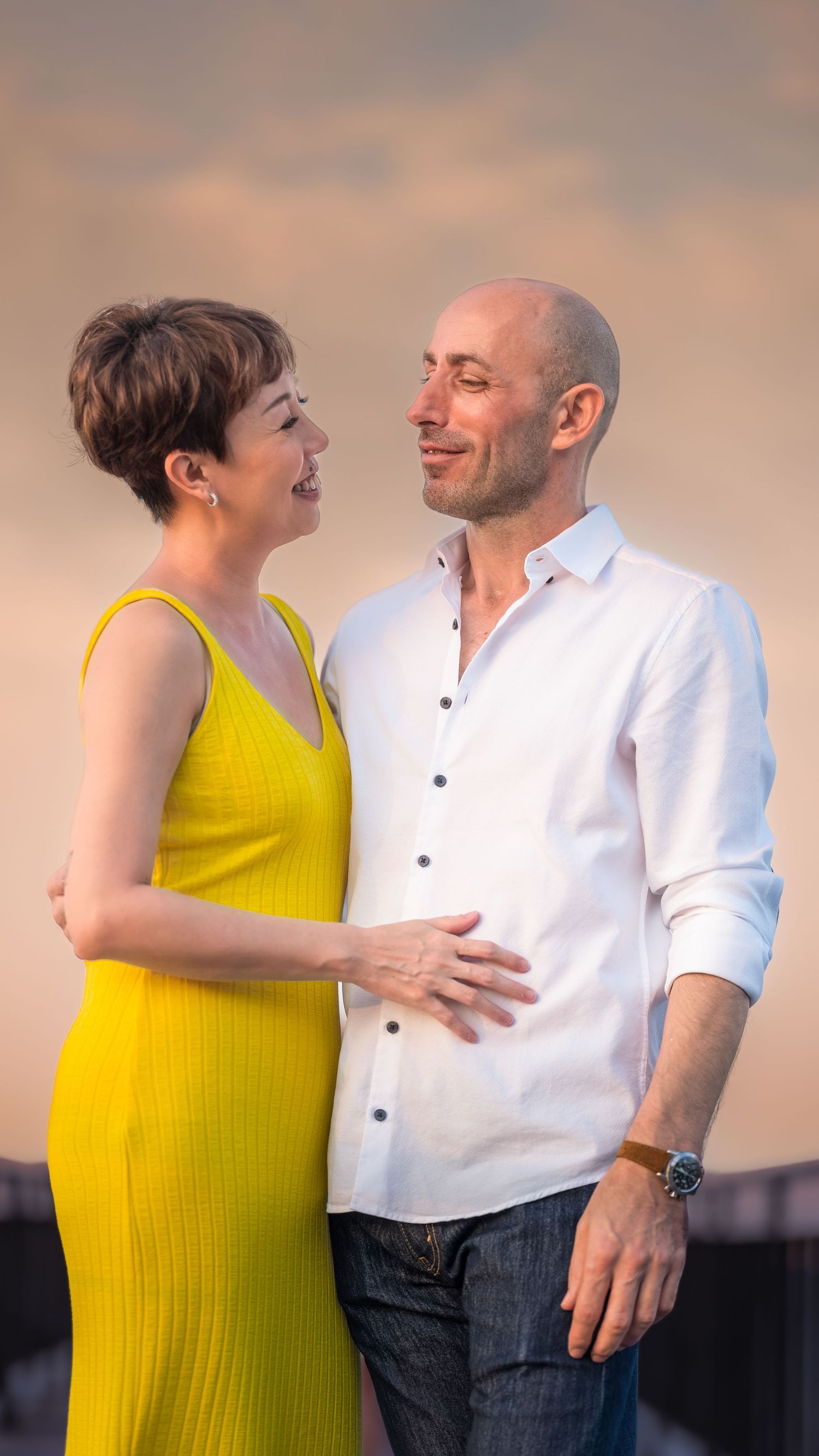 A couple in a tender moment, woman in yellow dress and bald man in white shirt smiling at each other at sunset.