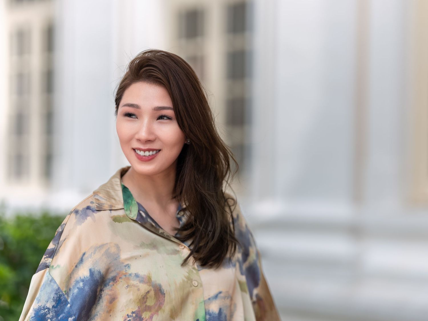 A smiling young woman with long brown hair wears a colorful floral wrap outdoors near a building.