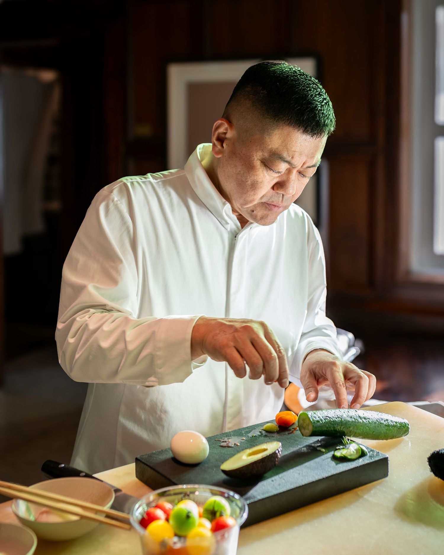 A chef in a white jacket carefully slices fresh vegetables on a dark cutting board in a warmly lit kitchen.