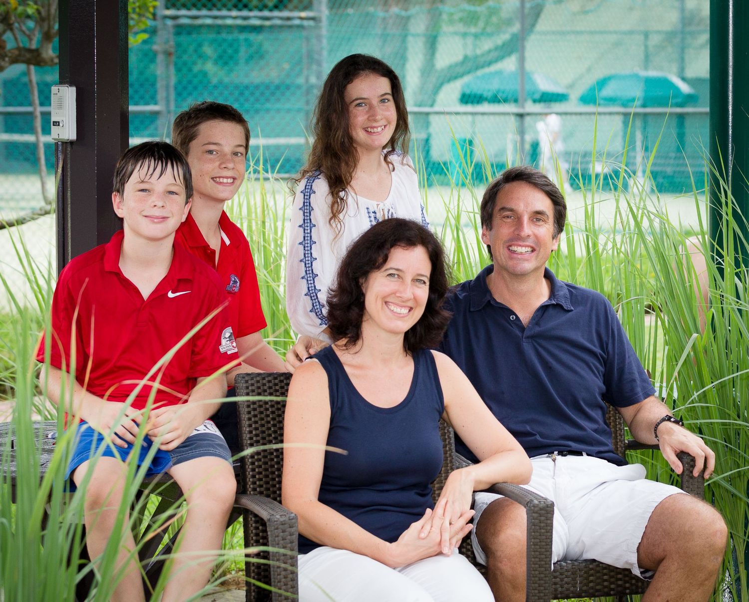 Happy family of four poses together on a patio near a tennis court in casual attire.