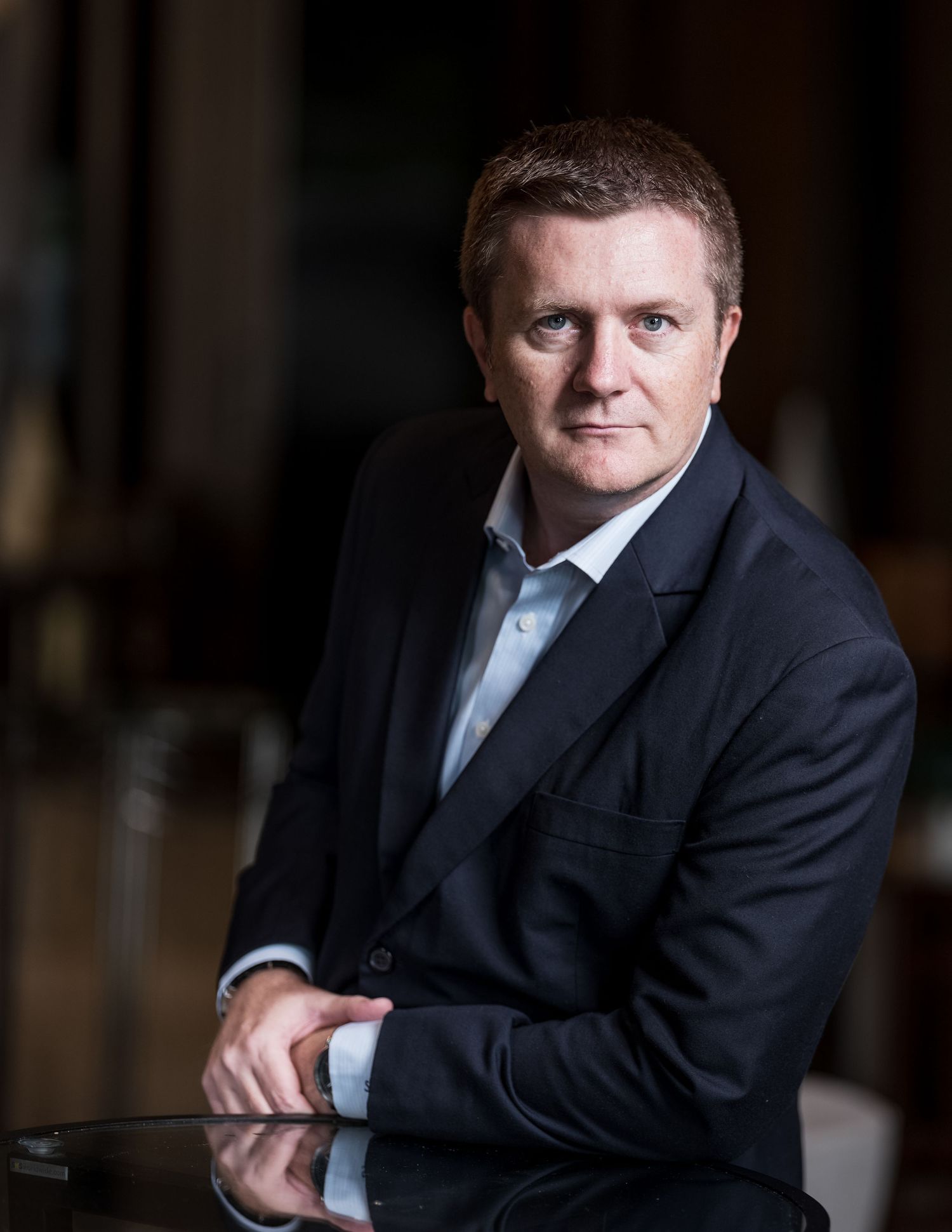 Professional man in dark blazer sits at a table in a dimly lit corporate portrait setting.