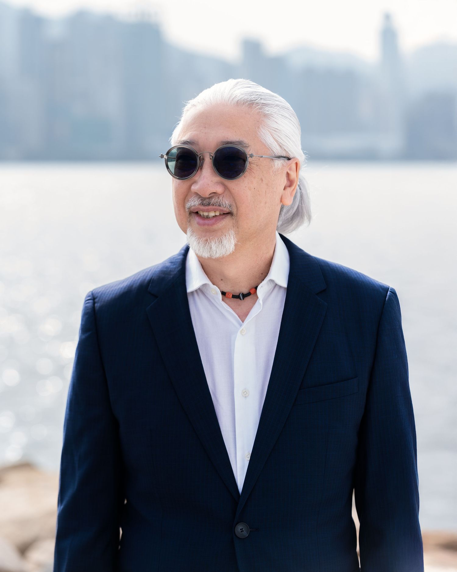 Stylish older man with gray hair and sunglasses wearing a navy blazer standing near a waterfront cityscape.
