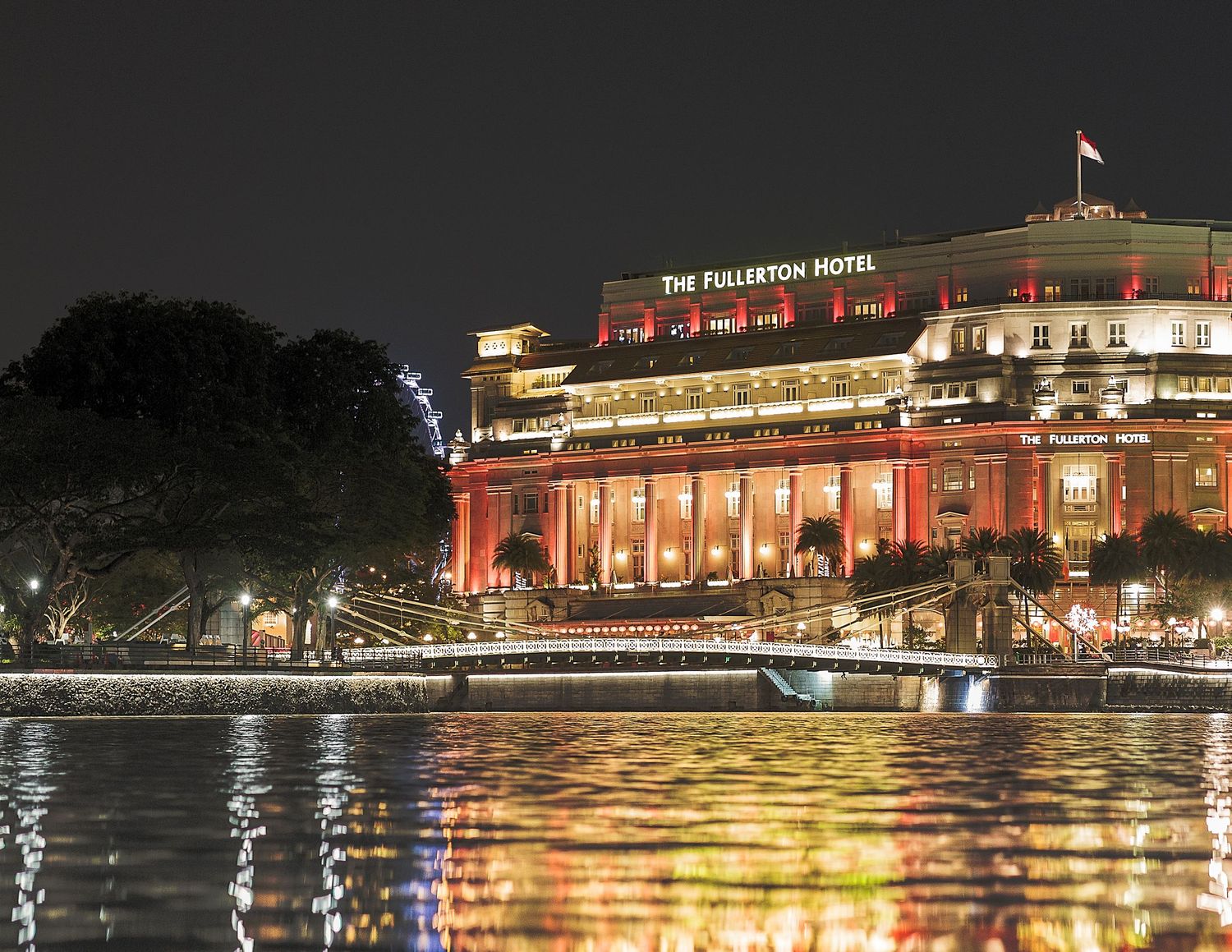 The Fullerton Hotel Singapore illuminated in red and gold lights reflected on the river at night.