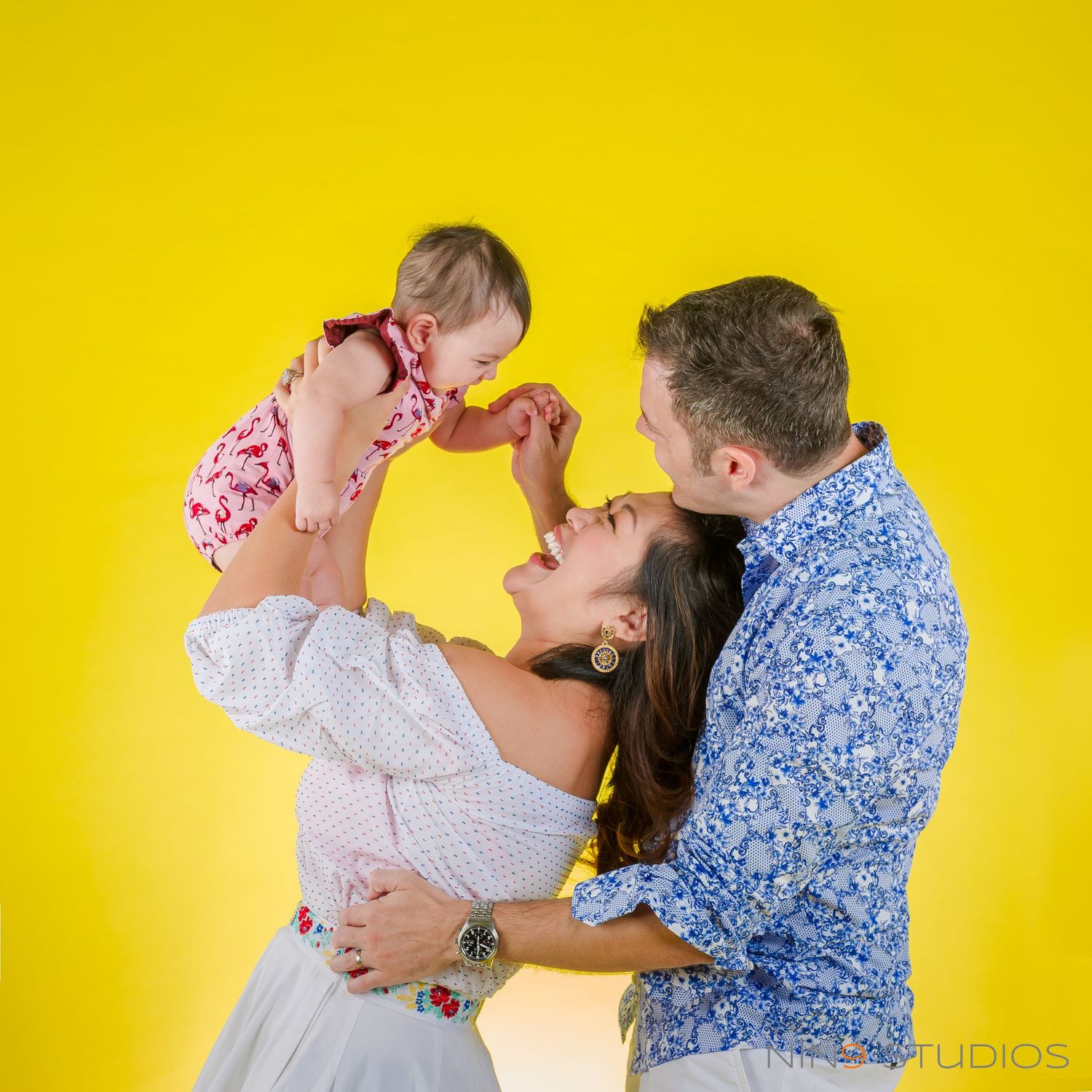 A joyful family poses against a yellow backdrop, with parents lifting their baby girl in a bright studio photo.
