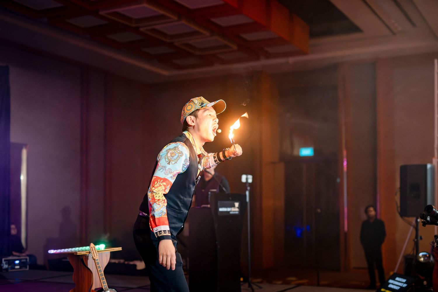 A tattooed performer breathes fire on stage at an indoor event venue with dramatic lighting.