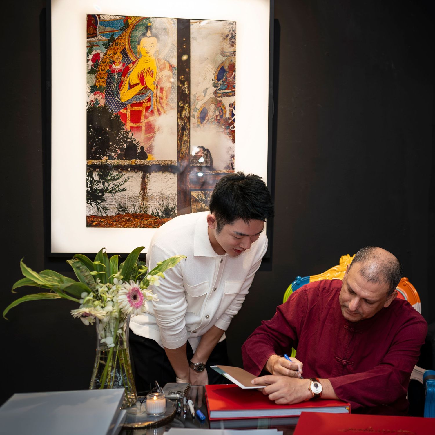 Man in red shirt signing a book at a desk beside a Buddhist artwork display in a gallery.