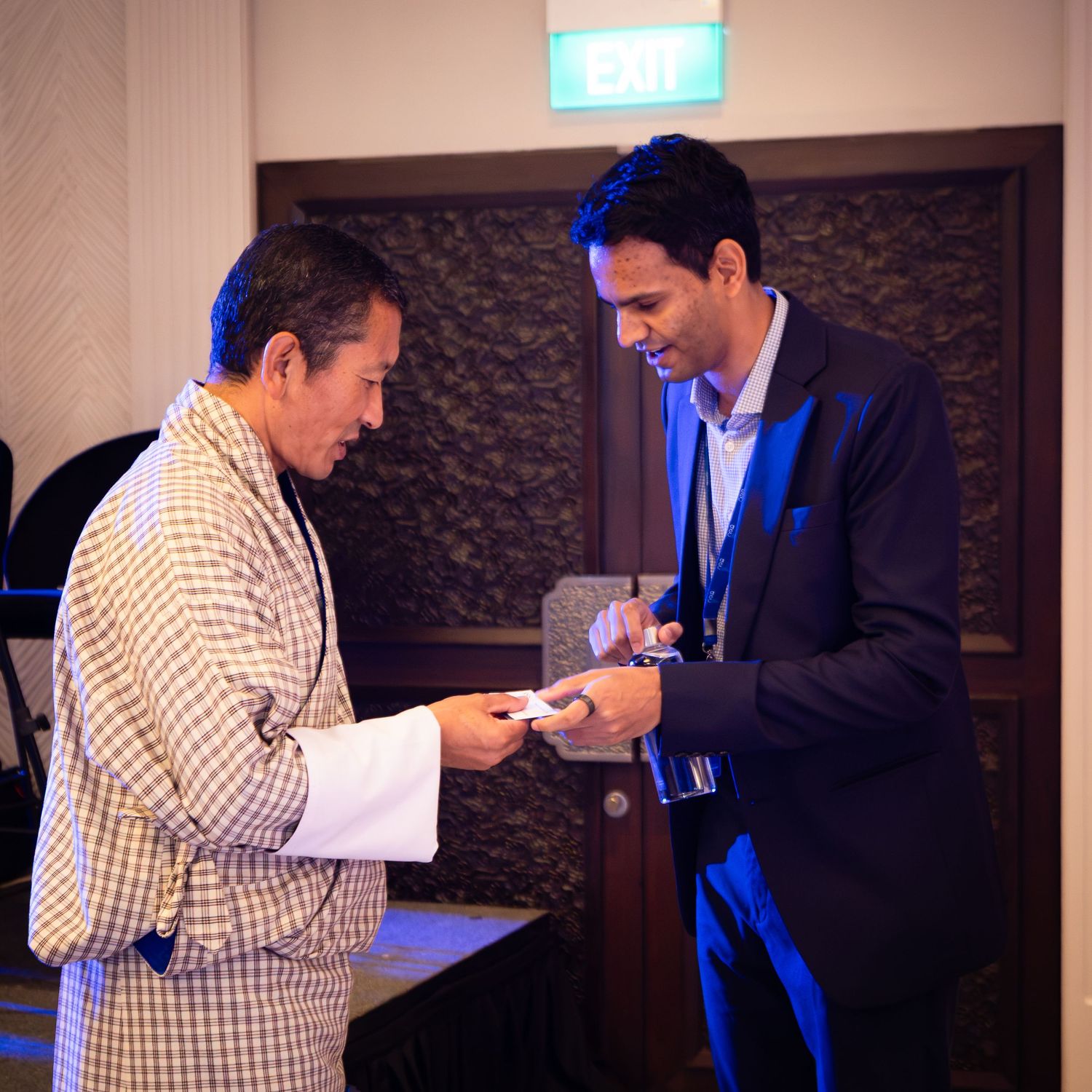 Two men exchange business cards in a dimly lit conference room setting.