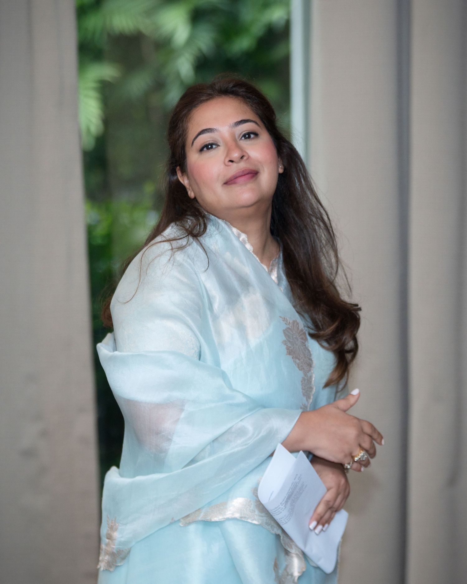Woman in light blue traditional Indian saree posing elegantly near a window with green foliage background.