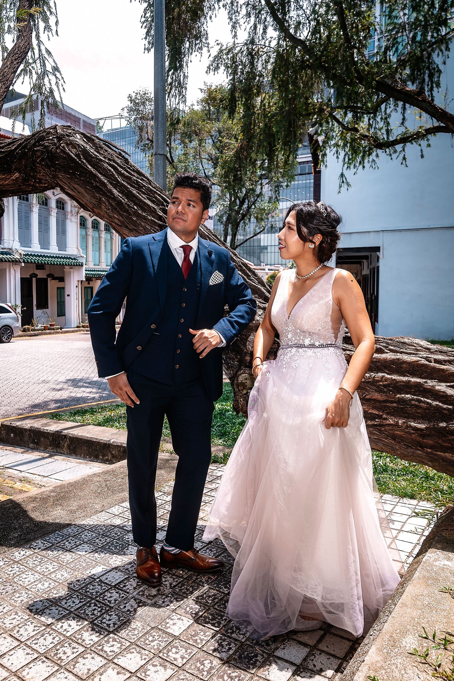 Couple in wedding attire posing outdoors, groom in navy suit and bride in white gown near a tree.