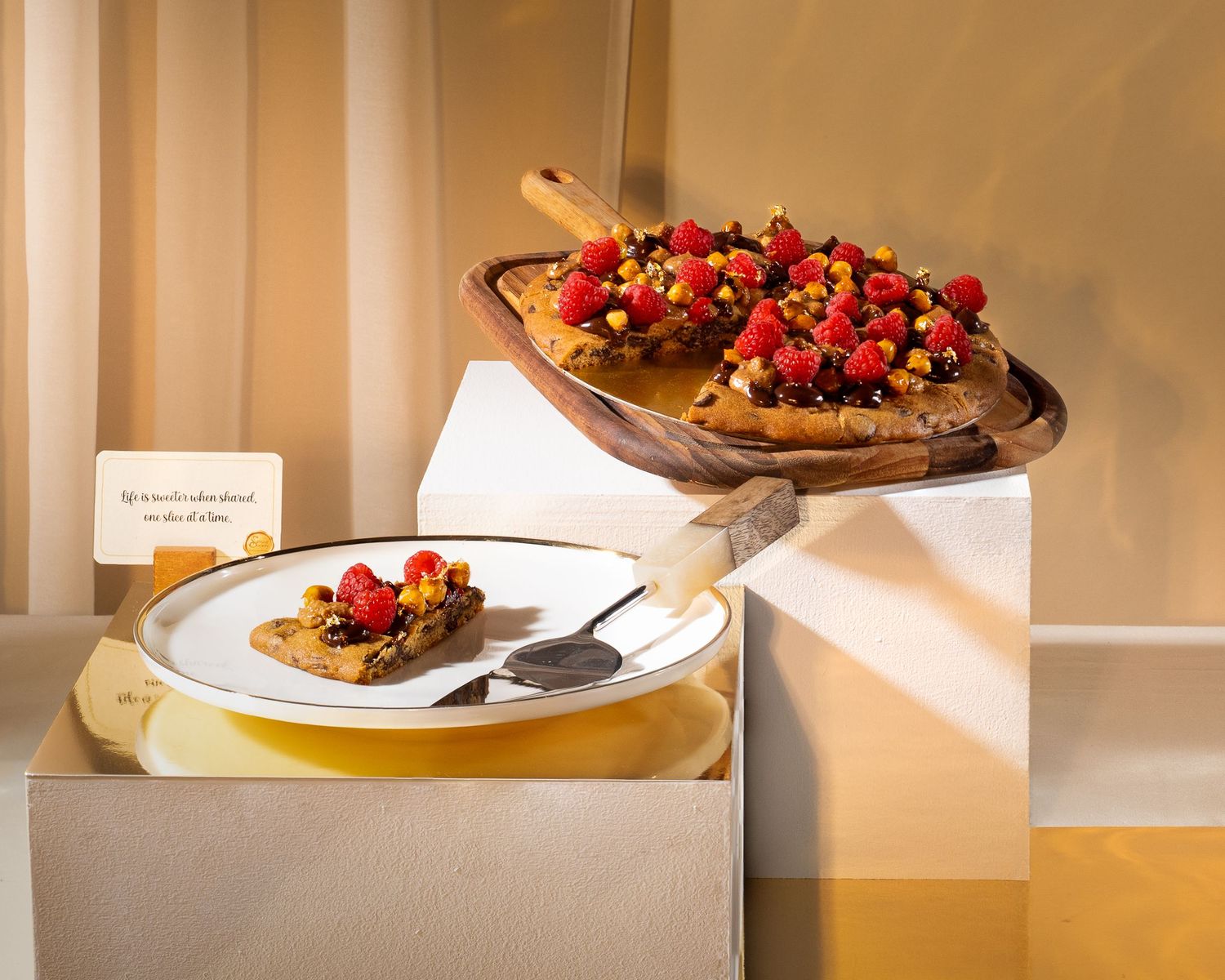 Elegant dessert display featuring raspberry-topped tarts on wooden board and white plate with spoon on geometric white pedestals.