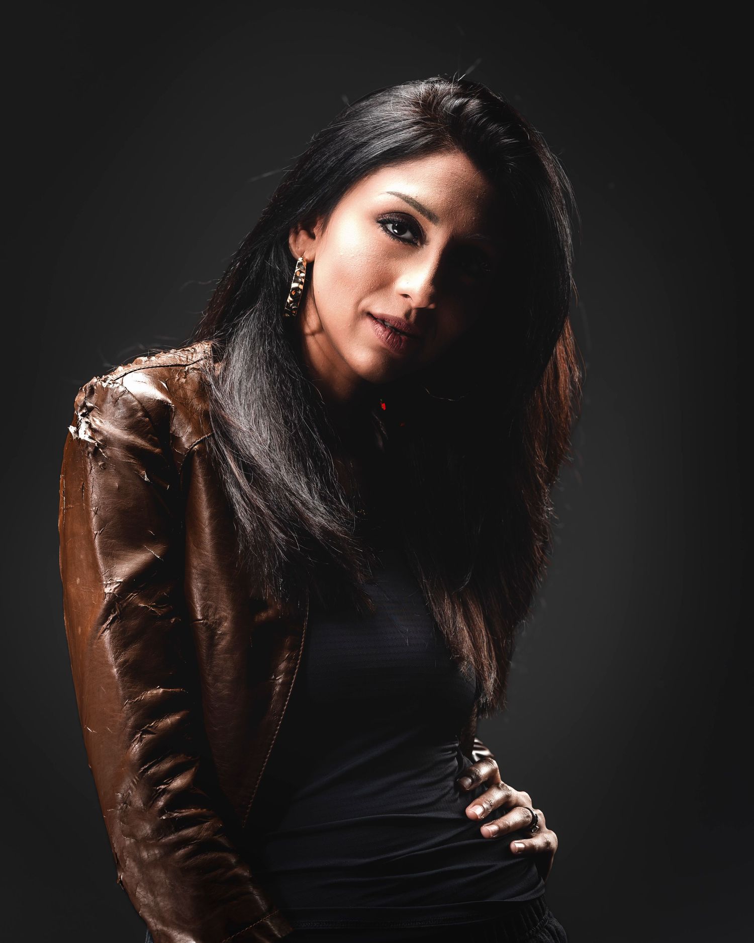 Woman in brown leather jacket and black top poses confidently against dark studio background with long black hair.