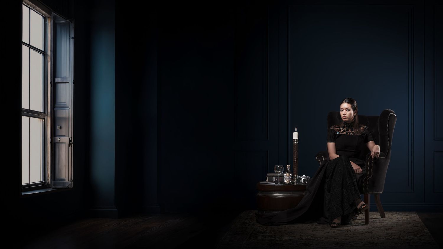 Woman in black dress seated in dark blue room with candle and window, gothic aesthetic portrait.