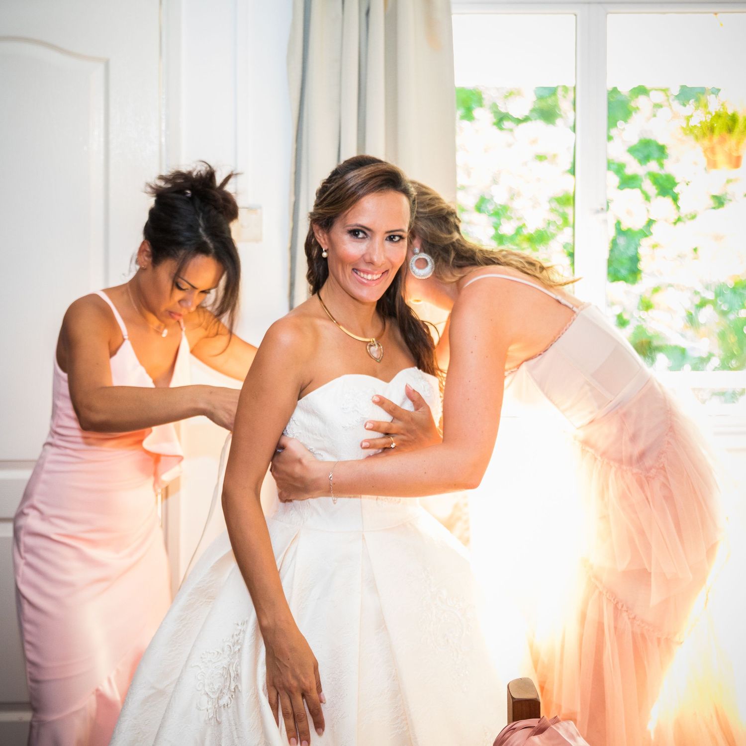 Bridesmaids helping a smiling bride in a white wedding gown get dressed before her ceremony.